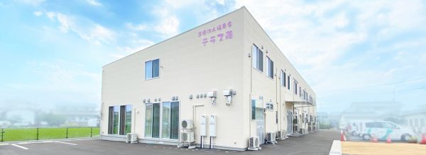 白い2階建ての介護施設の建物外観。駐車場と周辺環境が見える。